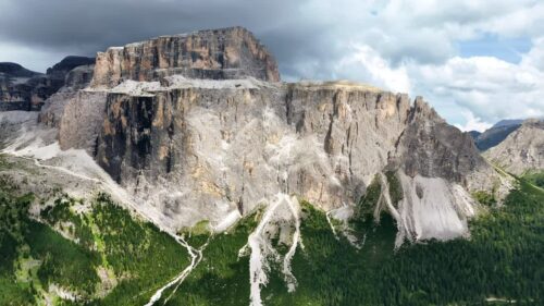 Video - Aerial drone view of a massive vertical limestone cliff rising above dense alpine forest. The imposing rock face dominates the mountainous landscape of the Dolomites