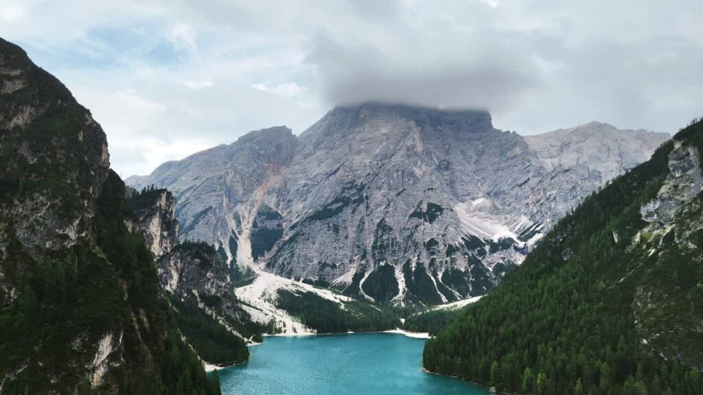 Video - Aerial drone view of a turquoise alpine lake surrounded by steep rocky mountains and dense pine forests. Clouds partially cover the high peaks of the Dolomites