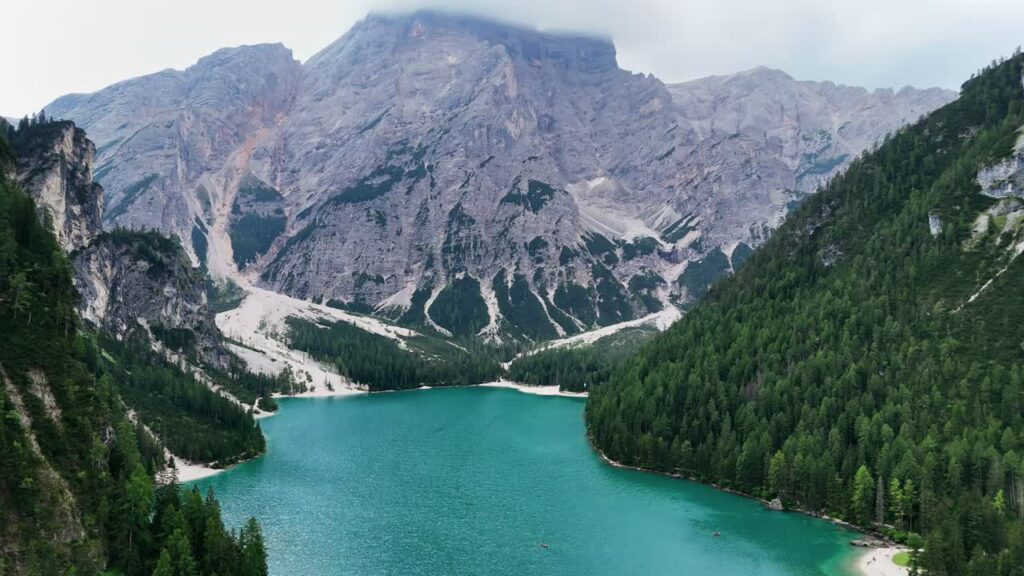 Video - Aerial drone view of the turquoise waters of Lago di Braies surrounded by dense alpine forest and dramatic limestone mountains in the Dolomites, Italy