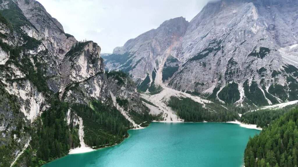 Video - Aerial drone view of the turquoise waters of Lago di Braies surrounded by dense alpine forest and dramatic limestone mountains in the Dolomites, Italy