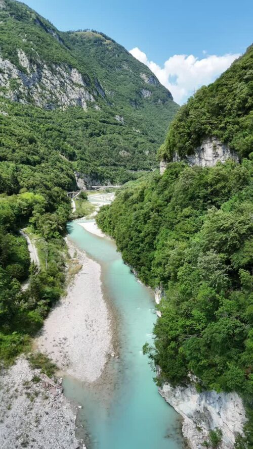 Video - Aerial drone view of a turquoise alpine river flowing through a lush green mountain valley with steep cliffs and dense forest. A small bridge crosses the valley in the distance as the river winds through the rugged landscape. Vertical