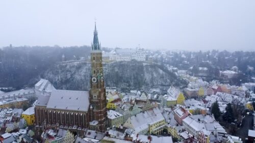 Video - Aerial drone view of Landshut Old Town covered in snow, featuring the tall Gothic tower of St. Martin's Church and Trausnitz Castle on the hill in Bavaria, Germany