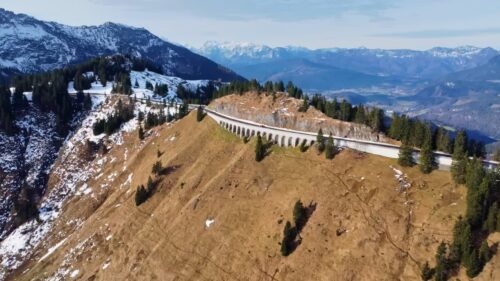 Video - Aerial drone view of a historic mountain road viaduct crossing a ridge in the Bavarian Alps. The curved stone structure stretches across a steep alpine slope with snow covered peaks in the background