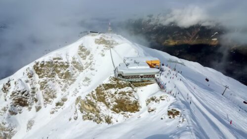 Video - Aerial drone view of a snow covered ski resort on a mountain summit with cable cars, ski lifts, and skiers on the slopes. Clouds drift across the dramatic alpine landscape in winter
