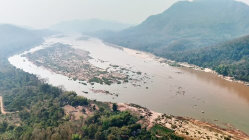 Video - Aerial drone view of the Mekong River flowing through mountains and forests in Laos, with sandbanks and islands visible in the wide river channel. Scenic Southeast Asian landscape with misty hills and natural wilderness