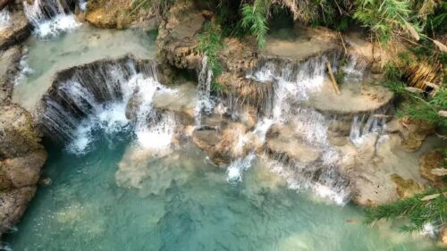 Video - Aerial drone view of turquoise pools and cascading limestone waterfalls at Kuang Si Falls, flowing through rock terraces and jungle vegetation in northern Laos