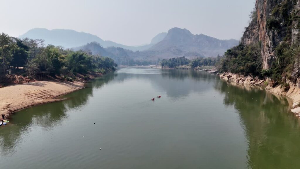 Video - Aerial drone view of two kayakers paddling on a calm river surrounded by limestone mountains and jungle landscape in Laos, creating a peaceful tropical travel scene