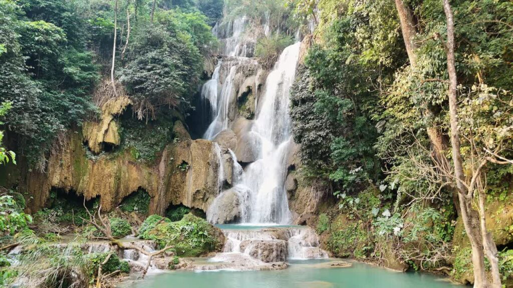 Video - Aerial drone view of Kuang Si Waterfall main cascade near Luang Prabang, where multiple streams of water fall down limestone rock formations into turquoise pools surrounded by dense jungle