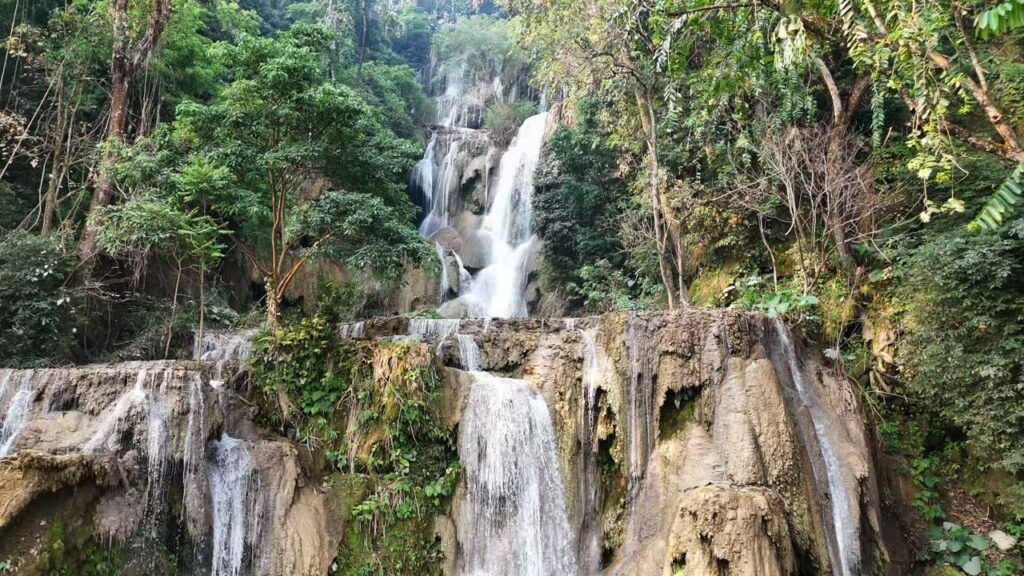 Video - Aerial drone view of Kuang Si Waterfall main cascade near Luang Prabang, where multiple streams of water fall down limestone rock formations into turquoise pools surrounded by dense jungle