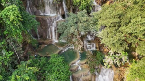 Video - Aerial drone view of Kuang Si Waterfall main cascade near Luang Prabang, where multiple streams of water fall down limestone rock formations into turquoise pools surrounded by dense jungle