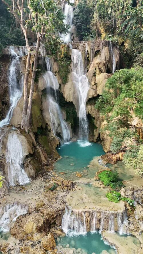 Video - Aerial drone view of multi-level turquoise pools and cascading waterfalls at Kuang Si Falls, surrounded by lush tropical forest in northern Laos. Vertical
