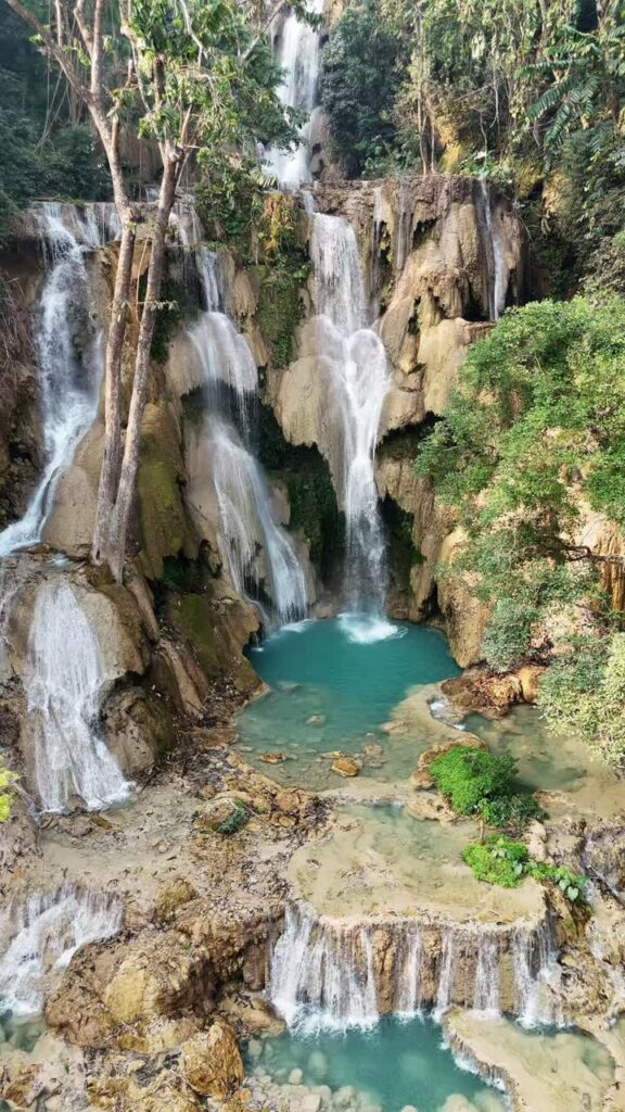 Video - Aerial drone view of multi-level turquoise pools and cascading waterfalls at Kuang Si Falls, surrounded by lush tropical forest in northern Laos. Vertical