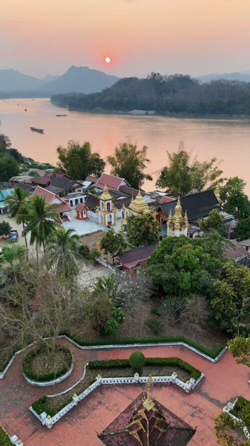 Video - Aerial drone view overlooking Luang Prabang temples and rooftops at sunset, with the Mekong River flowing through the valley and mountains in the background. Golden evening light reflects on the river as the sun sets over northern Laos. Vertical