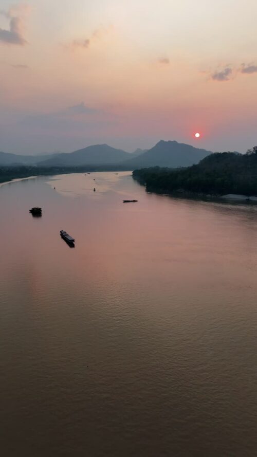 Video - Aerial drone view of boats traveling along the Mekong River during sunset in Laos, with warm orange light reflecting on the calm water and mountains fading into the distance. Vertical