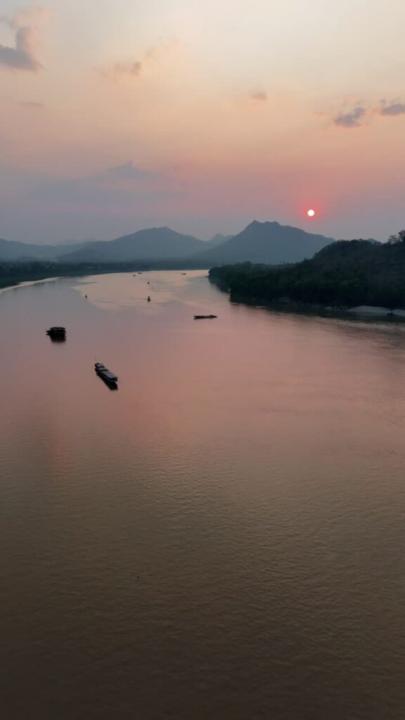 Video - Aerial drone view of boats traveling along the Mekong River during sunset in Laos, with warm orange light reflecting on the calm water and mountains fading into the distance. Vertical