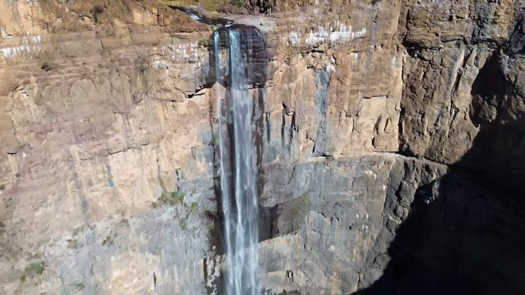 Video - Aerial drone view of Maletsunyane Falls in Lesotho, a powerful waterfall plunging down a high sandstone cliff with a rainbow forming in the mist below