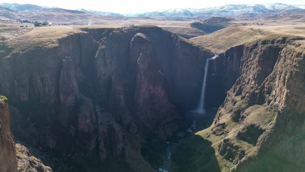 Video - Aerial drone view of Maletsunyane Falls dropping into a deep canyon in the Lesotho highlands, surrounded by rugged cliffs and vast mountain landscapes