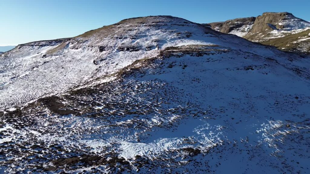 Video - Aerial drone view of snow covering mountain slopes in the Lesotho highlands, with rugged terrain and winter landscape under clear blue skies