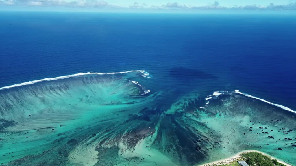 Video - Aerial drone view of turquoise lagoon waters and coral reef formations in Mauritius, with waves breaking along the reef line in the Indian Ocean