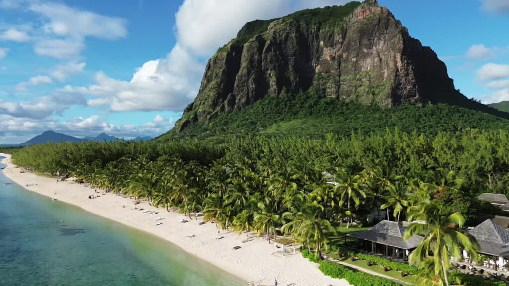 Video - Aerial drone view along a white sandy beach lined with palm trees at the base of Le Morne Brabant mountain in Mauritius, with turquoise lagoon water and a tropical resort along the shoreline