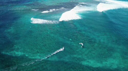 Video - Aerial drone view of a kitesurfer gliding across turquoise water above coral reefs in Mauritius, leaving a trail behind while waves break along the reef line