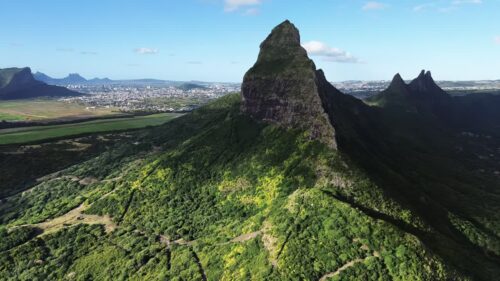 Video - Aerial drone view of a dramatic volcanic mountain peak surrounded by green hills with the city landscape visible in the distance in Mauritius
