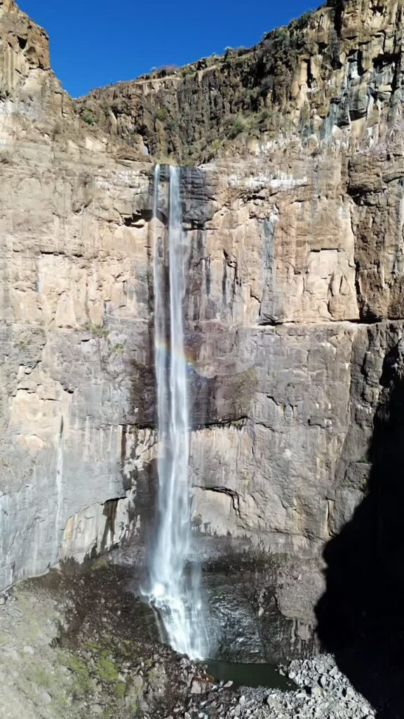Video - Aerial drone view of a tall waterfall dropping from a rocky cliff into a canyon below, with a faint rainbow visible in the mist and rugged mountain landscape surrounding it. Vertical