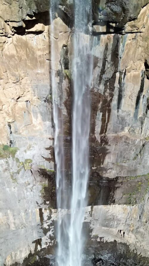 Video - Aerial drone view of a tall waterfall dropping from a rocky cliff into a canyon below, with a faint rainbow visible in the mist and rugged mountain landscape surrounding it. Vertical