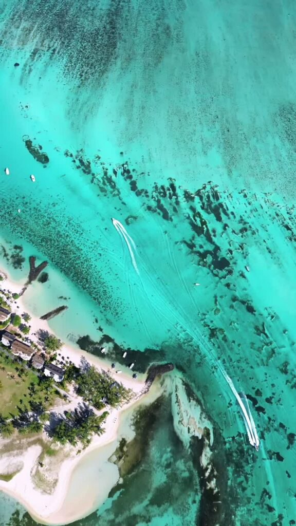 Video - Aerial drone view of a bright turquoise lagoon with speedboats moving through shallow water and coral patches near a sandy beach and tropical coastline. Vertical