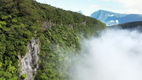 Video - Aerial drone view along a steep rainforest cliff with low clouds and mist rising from the valley, revealing dramatic mountains and untouched tropical wilderness in Madagascar