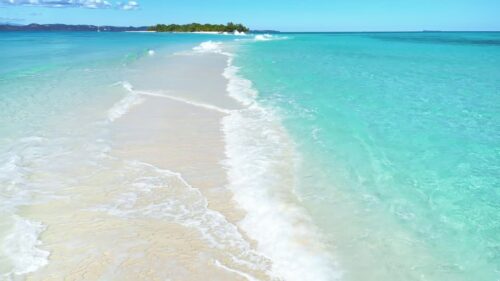 Video - Aerial drone view above a long white sandbar surrounded by crystal clear turquoise ocean water, leading toward a small tropical island in Madagascar