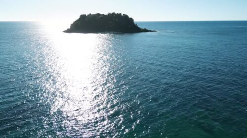 Video - Aerial drone view approaching a small forested island surrounded by deep blue ocean water, with sunlight reflecting on the sea surface off the coast of Madagascar