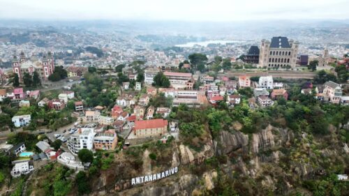 Video - Aerial drone view above Antananarivo with the city skyline spread across surrounding hills, showing dense urban neighborhoods and the large historic palace complex overlooking Madagascar's capital