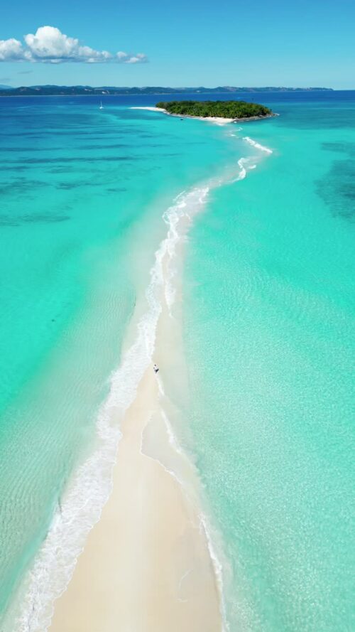 Video - Aerial drone view of a narrow white sandbar cutting through bright turquoise water, leading toward a small tropical island in Madagascar's Indian Ocean lagoon. Vertical