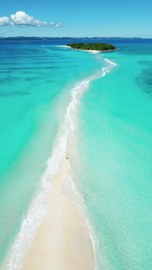 Video - Aerial drone view of a narrow white sandbar cutting through bright turquoise water, leading toward a small tropical island in Madagascar's Indian Ocean lagoon. Vertical