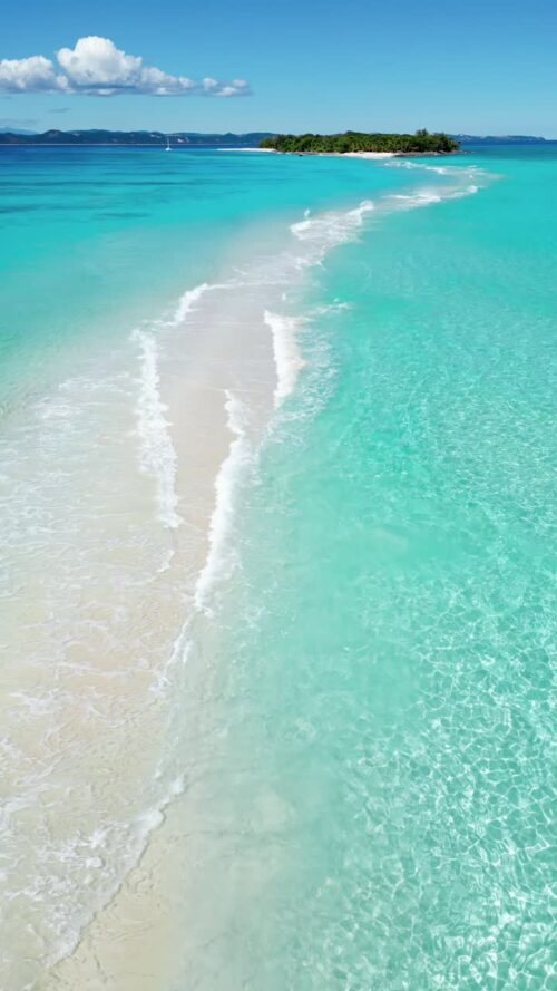 Video - Aerial drone view of a narrow white sandbar cutting through bright turquoise water, leading toward a small tropical island in Madagascar's Indian Ocean lagoon. Vertical
