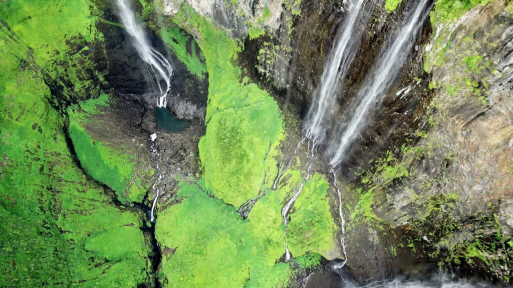 Video - Aerial drone view of multiple narrow waterfalls flowing down steep green cliffs, surrounded by dense vegetation in a tropical mountain landscape