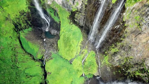 Video - Aerial drone view of multiple narrow waterfalls flowing down steep green cliffs, surrounded by dense vegetation in a tropical mountain landscape