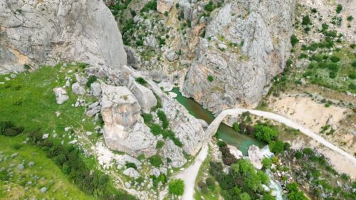 Video - Aerial drone view of rocky mountains and canyon landscapes with a small stone bridge crossing a river below, surrounded by rugged terrain and green valley vegetation in Mesopotamia