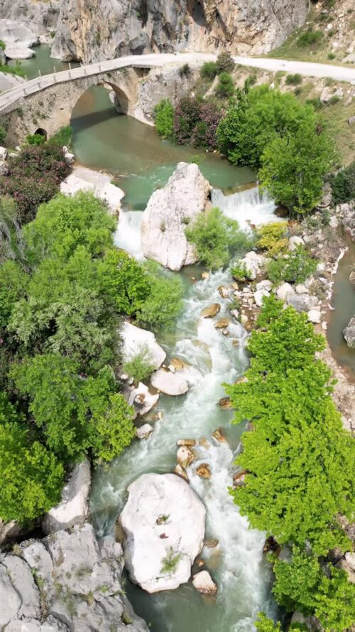 Video - Aerial drone view of a mountain river flowing under a small bridge surrounded by rocks and green trees in a canyon valley in Mesopotamia. Vertical