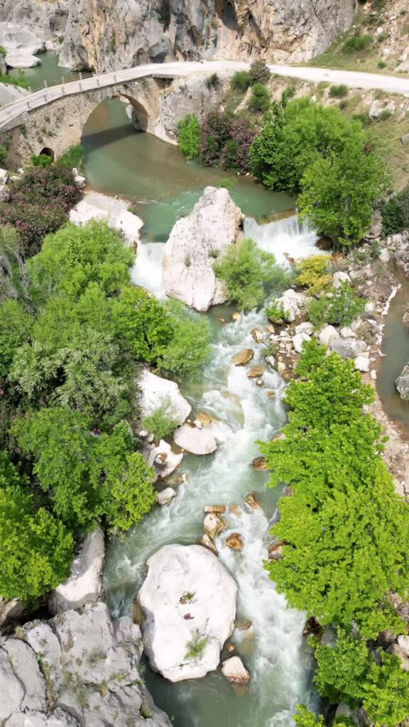 Video - Aerial drone view of a mountain river flowing under a small bridge surrounded by rocks and green trees in a canyon valley in Mesopotamia. Vertical
