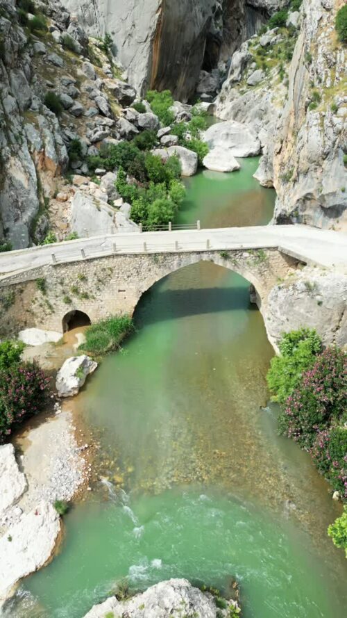 Video - Aerial drone view of a mountain river flowing under a small bridge surrounded by rocks and green trees in a canyon valley in Mesopotamia. Vertical