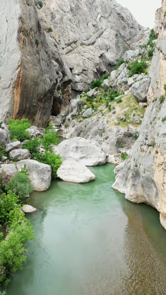 Video - Aerial drone view of a mountain river flowing under a small bridge surrounded by rocks and green trees in a canyon valley in Mesopotamia. Vertical