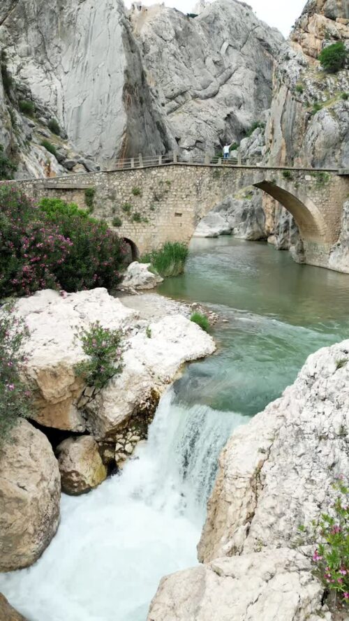 Video - Aerial drone view showing a small waterfall flowing beneath a historic stone bridge in a rocky canyon landscape of Madagascar. Vertical