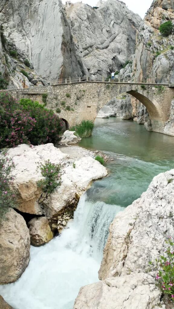 Video - Aerial drone view showing a small waterfall flowing beneath a historic stone bridge in a rocky canyon landscape of Madagascar. Vertical