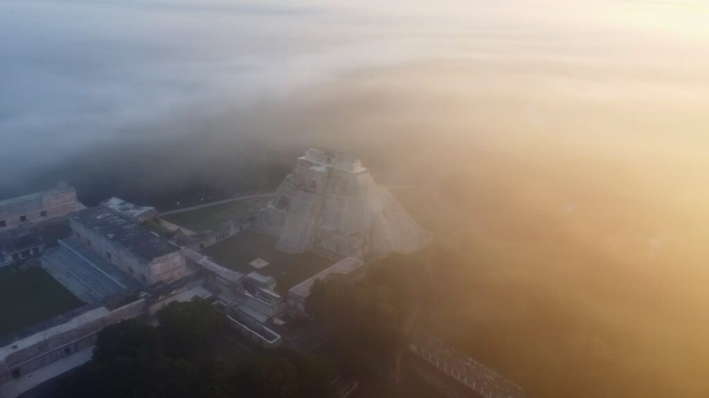 Video - Aerial drone view of the Temple of the Inscriptions at Palenque archaeological site emerging through morning mist in the Chiapas jungle, Mexico, showing ancient Mayan ruins surrounded by dense rainforest