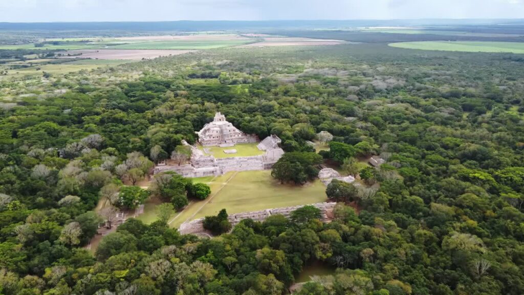 Video - Aerial drone view of Edzna Mayan pyramid rising above dense tropical forest in Campeche, Mexico, revealing ancient Mesoamerican ruins surrounded by jungle landscape