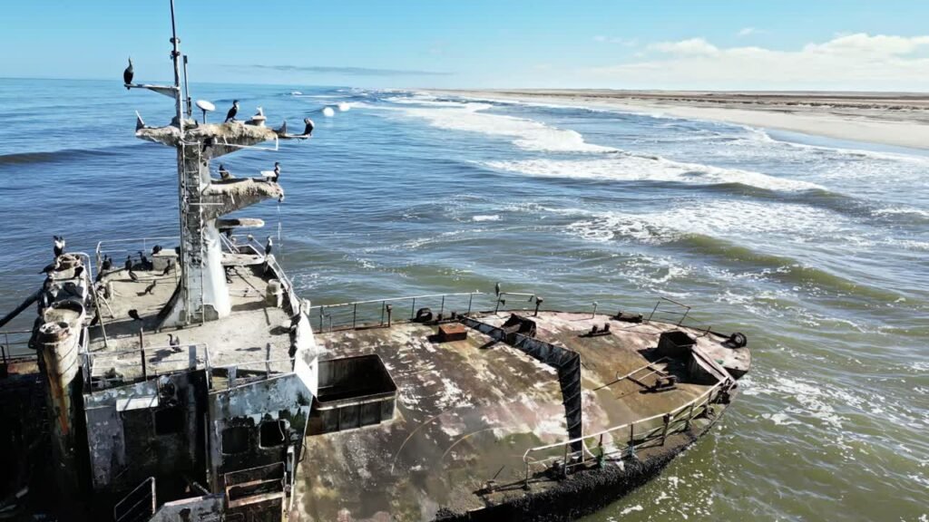 Video - Aerial drone view of an abandoned shipwreck on the remote Skeleton Coast of Namibia with ocean waves breaking along the sandy shoreline and seabirds resting on the rusted vessel