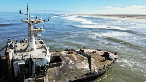 Video - Aerial drone view of an abandoned shipwreck on the remote Skeleton Coast of Namibia with ocean waves breaking along the sandy shoreline and seabirds resting on the rusted vessel