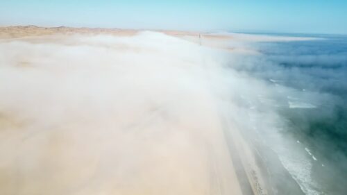 Video - Aerial drone view of coastal fog moving over vast desert dunes along the Atlantic Ocean in Namibia creating a dramatic atmospheric landscape